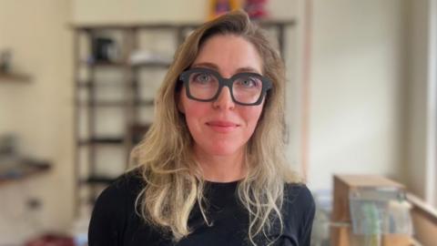 Claire Mulvenna stands in her opticians shop. She is wearing thick black rimmed glasses, and a a black jumper. She has long blond hair.