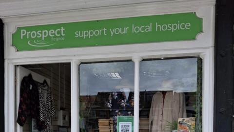 The top of a charity shop front with clothes and books in the window and a green sign that says 'Prospect Hospice - support your local hospice'
