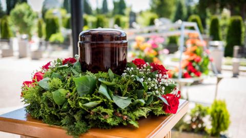 A flower wreath around a urn sitting on a table. In the background there are other flowers on a metal structure.