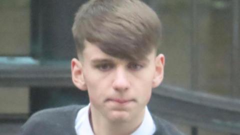 A young man with brown hair that is styled into a fringe, wearing a shirt and grey jumper.