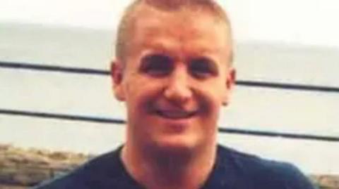 A picture of a smiling Joseph Cummins, with short blond hair and wearing a blue T-shirt, with a view of the sea visible in the background.