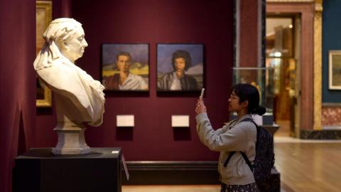 A young woman with black hair, glasses and a backpack stands in front of a statue of a bust and takes a photo. Portraits line the wall behind her.