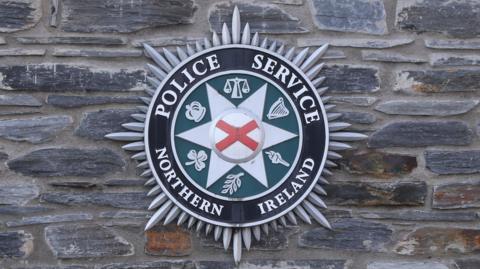 On a grey stone wall hangs a PSNI badge. The badge is silver black and green