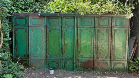 The seven tall green iron panels have rusted over time. The panels make up one side of the shelter and are surrounded by greenery.