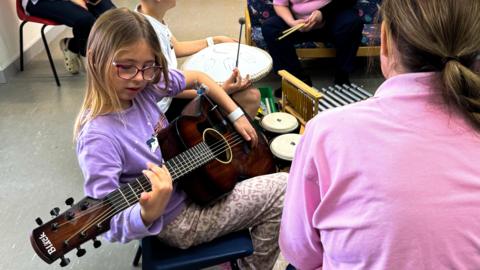 A young girl in mauve pyjamas plays a guitar with a woman in a pick top looking on