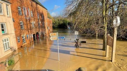 Brown floodwater submerges a road and path a street sign stand in the centre with brick buildings to the left reflecting in the water.