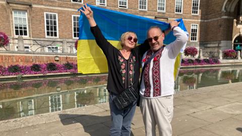 A woman and a man arm in arm, holding up a blue and yellow Ukrainian flag behind them as they stand on Bristol's College Green. They are both wearing Ukrainian-style embroidered clothes.