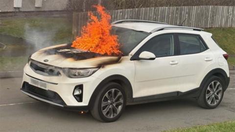 The empty white Kia car stopped in the middle of a road by a house with a wooden fence, its engine on fire with flames licking the windscreen and charring on the bonnet