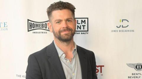 Jack Osbourne, who has dark hair cropped short and a beard, standing in front of a hoarding bearing various company logos. He is wearing a grey suit jacket and pale grey shirt and smiling.
