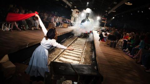 A steam train in the background, with a girl in the foreground holding a red flag. The train is in a theatre, with the audience sitting on either side of the track.