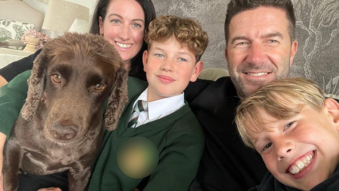 A woman and man, both with dark hair, sat on a sofa in front of a grey wallpaper backdrop. In front of them are two blonde boys, one with curly hair and the other with straight, and a brown cocker spaniel. All four people are looking at the camera and smiling.