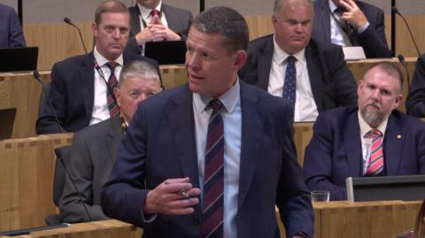 Rhun ap Iorwerth speaking in the Senedd, surrounded by other Members of the Senedd.