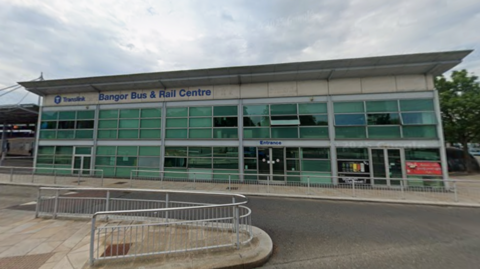 A building with lettering in blue stating: Translink. Bangor Bus & Rail Centre. The word entrance is displayed over a door. The sky is cloudy. 