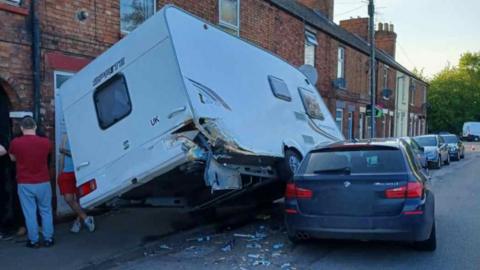 A caravan off its wheels with a black BMW lodged underneath it on a street with terraced brick housing
