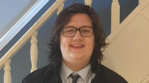 Wearing dark framed glasses, a white shirt, dark tie, and with shoulder length dark brown hair, Ethan Entwistle looks to the camera with an open-mouthed smile. Behind him are the bannisters of a flight of stairs.