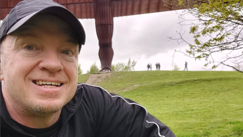 A man in a black baseball cap and black top smiling at the camera. He is outside and there is grass in the background.