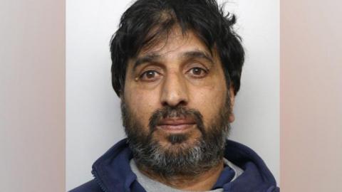 A police mugshot of Qabir Hussain. He has  a greying beard and black hair. He is  wearing a navy fleece over a grey top.