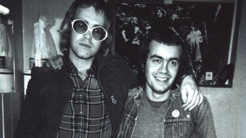 A black and white photo of Elton John wearing large glasses, with his arm around Bernie Taupin. Bernie is wearing a denim jacket over a jumper. Elton