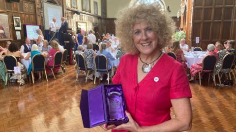 Ms Barlow wears a bright fusia dress and has blond curly hair in a bob.  She holds the glass award which sits in a purple satin box.  The volunteers sit around tables in the background within the wood panelled room of the Guildhall.