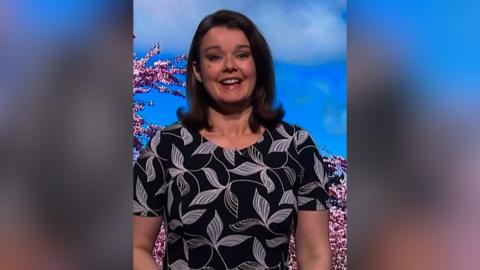 Gillian Smart smiles as she stands in front of a green screen which is showing a blue sky, clouds, and pink trees. She has brown shoulder-length hair and is wearing a black dress with a white leaf design printed on it.