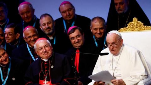 Pope Francis speaks next to Cardinal Jean-Marc Aveline, Archbishop of Marseille during a meeting at Palais du Pharo, on the occasion of the Mediterranean Meetings (MED 2023), in Marseille, France, September 23, 2023. There are eight other priests wearing vestments, while Francis is holdingRe pieces of paper and speaking into a micropghine.
