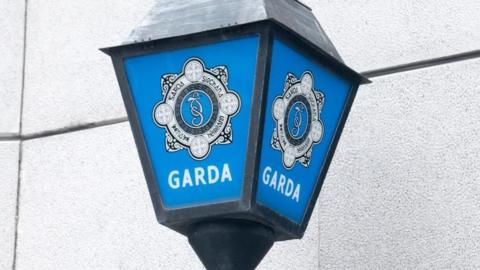 Blue and white police sign beside a grey building, featuring crest and the word GARDA in block capitals.