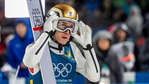 Daniel Tschofenig holds his hands up to his goggles and is wearing a gold helmet with a pair of skis on his shoulder