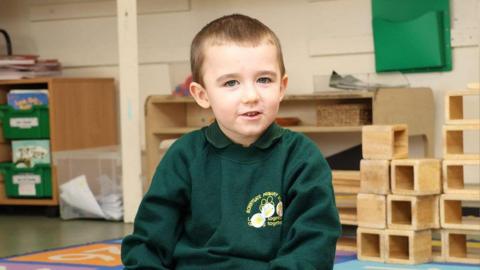 A young boy dressed in a green school sweatshirt and polo shirt has a half smile on his face. The background shows school equipment and trays.