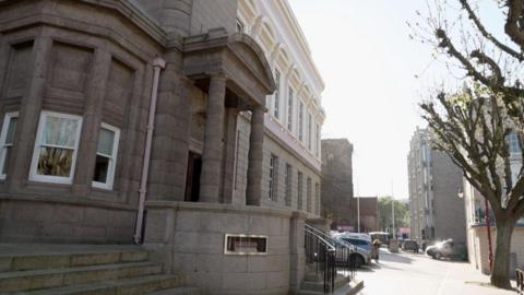 A stone building with a number of windows. A path runs in front of the building. Cars are parked at the entrance. Small steps lead up to the entrance of the building.