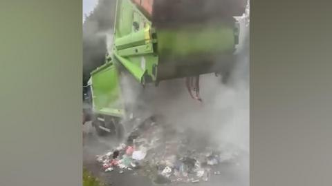 A green bin lorry is partially opened up as waste is deposited on the road side with smoke pouring from the vehicle.