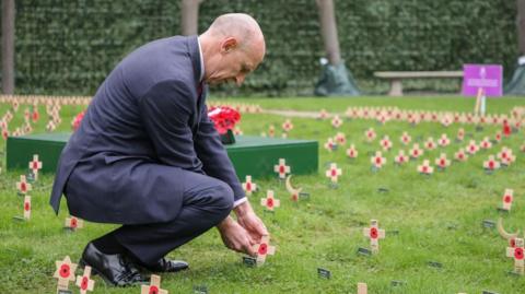 John Healey, wearing a suit, is planting a cross with a red poppy on it into the grass. Around him are many similar crosses, planted by other people.