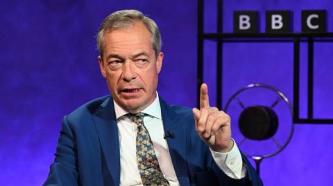 Nigel Farage holds up his finger, with a 91ȱ sign in the background