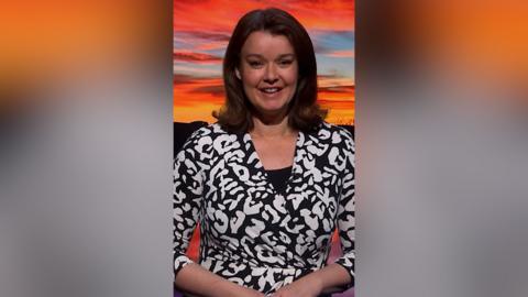 Weather presenter Gillian Smart smiles at the camera. She has mid-length brown hair and is wearing a black and white patterned dress. She is standing in front of a green screen that is showing a picture of Scotland.