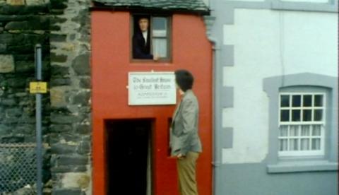 Britain's tallest mobile man in 1975 standing next to Britain's smallest house. He towers over the door, able to easily reach the window on the top floor