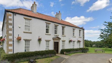 Shotton Hall. It is a two-storey white country house, surrounded by flowers and shrubs.
