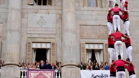 Acrobats in red tops and white pants stand in a human pyramid while people stand on a nearby balcony watching and clapping. 