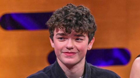 Owen Cooper smiles during Graham Norton interview. He has a mop of dark curly hair and wears a dark shirt. The backdrop is an orange wall decorated with purple lozenge shapes. It is a head shot of him. 