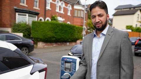 A man with black hair and beard, wearing a blue shirt and grey jacket, stands by a residential street and next to an electric vehicle charging point and the rear of a white car.