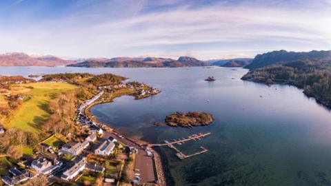 An aerial view of Plockton showing white-walled houses, the marina and Loch Carron. There hills and mountains in the distance.