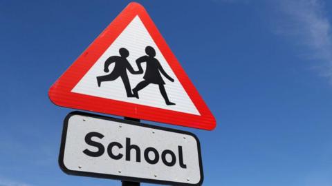 A road sign set against a blue sky. The sign shows the shadow of two children holding hands and walking on a white background, surrounded by a red triangle. The sign underneath reads 'school' in black font on a white background.