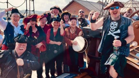 The Auckland Shanty Singers, with leader Christopher Miller (bottom left), cheering and dressed as pirates. There is a group of 11 people, plus one dog, on an old style ship.
