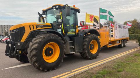 A yellow tractor with a trailer attached to the back. There are several flags attached to the back of the trailer.