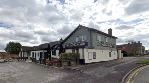 The pub stands on the left of the image, set back from the road with a wide pavement. There is a big car park to one side of the pub and some outdoor seating by some fencing. The pub has an external wood-effect on the top half of it and is painted a light grey/green colour. The bottom half is cream.