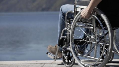 A stock close-up image of a person in a wheelchair showing a man's hand on a wheel rim and a foot in brown suede shoe on a footrest. The wheelchair is on a pavement overlooking a body of water.