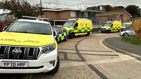 An ambulance car, a police car and two incident response vans parked up on the kerb of a residential street.