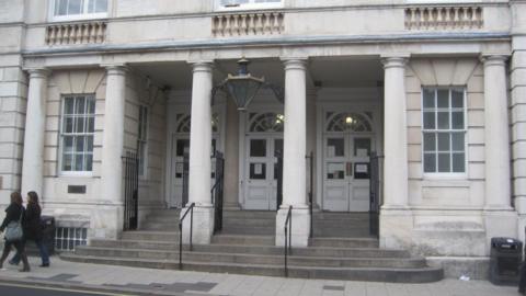 The exterior of a grey stone building. The image shows the steps and large pillar leading to three white doors.