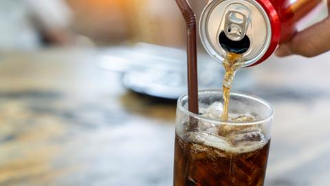Cola is poured from a red can into a glass with ice.