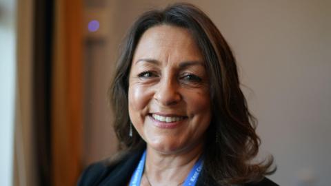 Linden Kemkaran, wearing a dark jacket and a Kent County Council lanyard, smiles into the camera in front of the background of a blurred room.