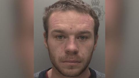 A police custody shot of Alex Fullard looking at the camera. He has short brown hair light brown facial stubble. He is wearing a grey top with a black neckline. The wall behind him is grey.