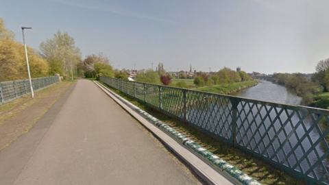 A tarmacked bridge that goes over a large river is pictured on a sunny day. There is a large patch of grass to the left of the river and the skyline of Hereford can be seen from the bridge. 